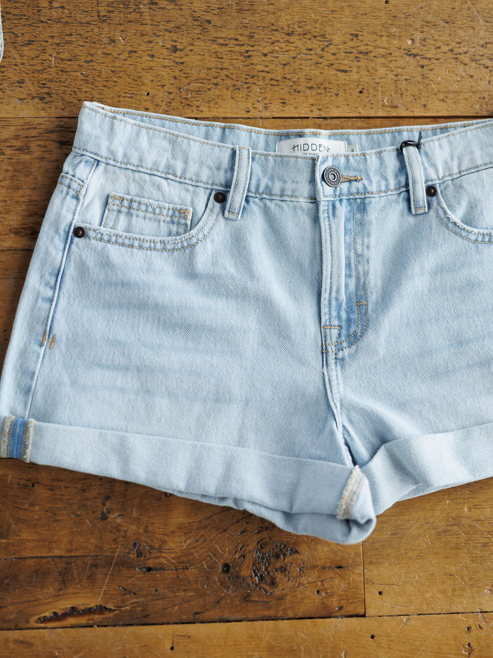 The Mackie Shorts by Savannah Sevens western life{&}style are light wash denim with rolled hems, front and coin pockets, belt loops, and a button-zipper closure, shown laid flat on a rustic wooden surface.