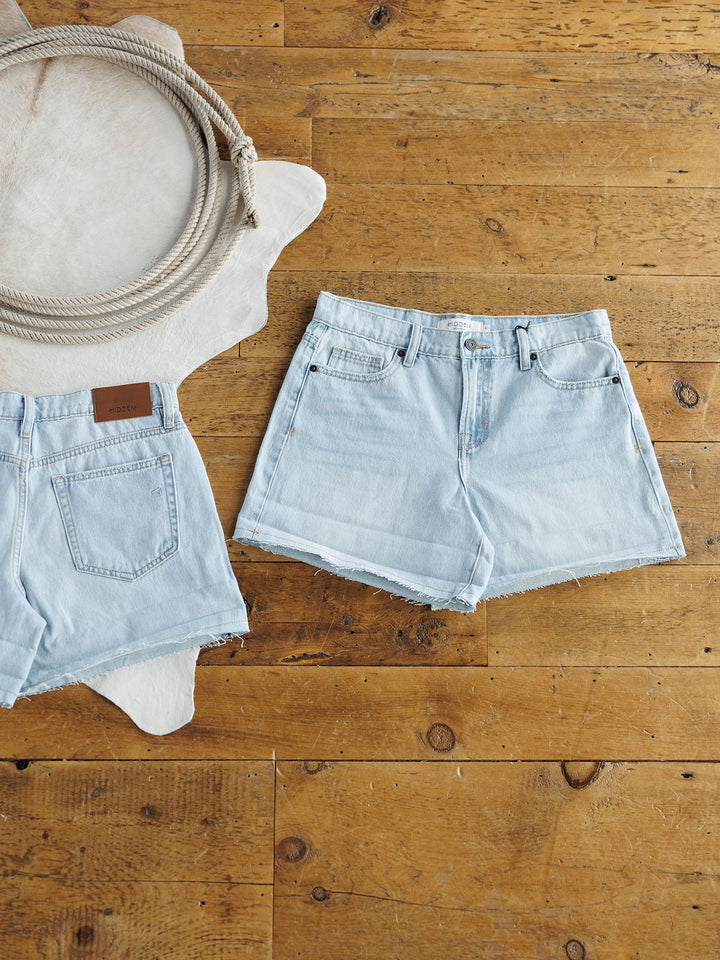 Two pairs of Savannah Sevens western life{&}style The Mackie Shorts are displayed on a wooden floor by a coiled rope and white cowhide rug—one pair shows a raw cut hem from the front, the other is shown from the back.