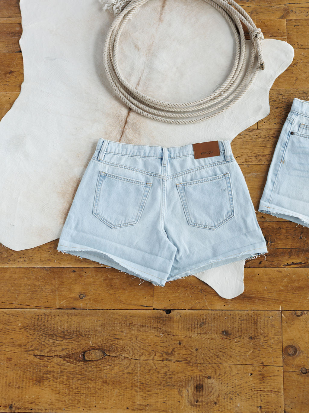 The Mackie Shorts by Savannah Sevens western life{&}style, light wash denim with frayed hems, are displayed on a white animal hide rug atop a wooden floor with a coiled rope placed above them.