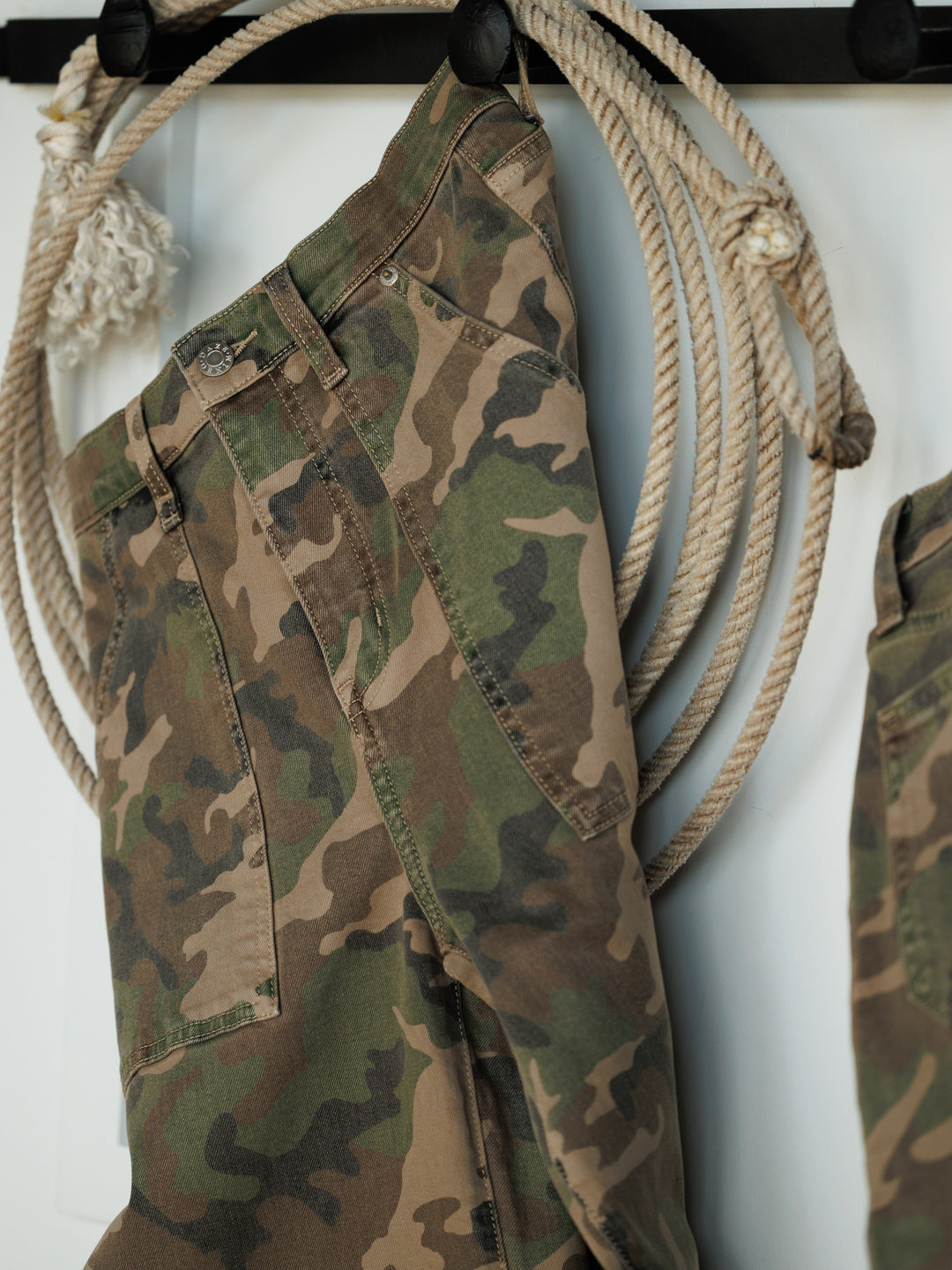 The Granger High Rise Wide Leg Jean by Savannah Sevens western life{&}style features a camo pattern and hangs on a hook, complemented by thick ropes. Its wide-leg cut stands out against the minimalist, neutral-toned backdrop.