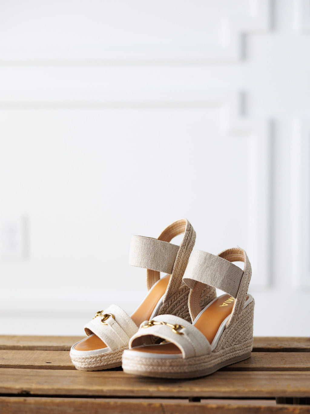 The Abilene by Savannah Sevens western life{&}style: Beige wedge sandals with open toes, thick ankle straps, and gold snaffle bit accents on the toe straps, shown on a wooden surface against a white textured wall.