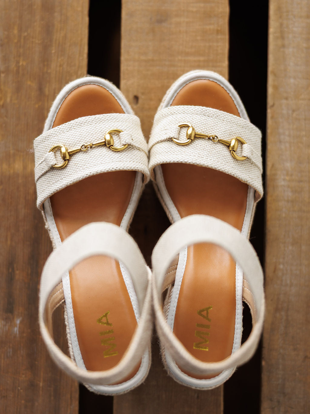 The Abilene by Savannah Sevens western life{&}style: tan wedge sandals with open toe and gold snaffle bit details on the straps, displayed on wood. The insoles feature "MIA" in gold letters.