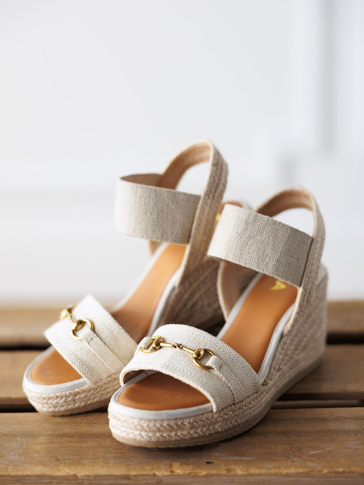 The Abilene by Savannah Sevens western life{&}style: beige open toe wedge sandals with ankle straps and gold snaffle bit accents, featuring woven espadrille-style soles, displayed on a wooden surface.