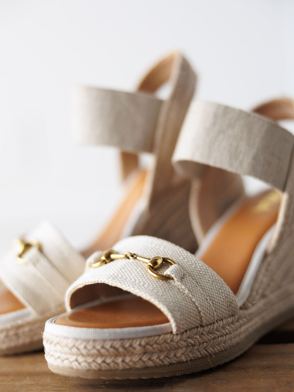 A close-up of The Abilene by Savannah Sevens western life{&}style—beige, open-toe wedge sandals with canvas straps and a gold snaffle bit detail, displayed on a light wooden surface.