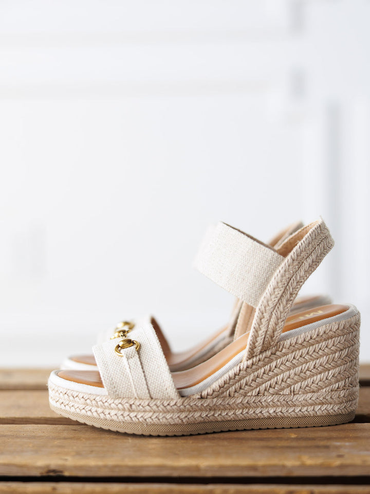 The Abilene by Savannah Sevens western life{&}style: a beige open-toe wedge sandal with braided espadrille sole, ankle strap, and gold chain detail on the front, displayed on a wooden surface against a white background.