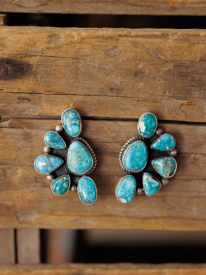 The Watseka Earrings by Savannah Sevens western life{&}style are handcrafted Native American sterling silver earrings with multiple turquoise stones in a circular floral design, shown on a rustic wooden background.