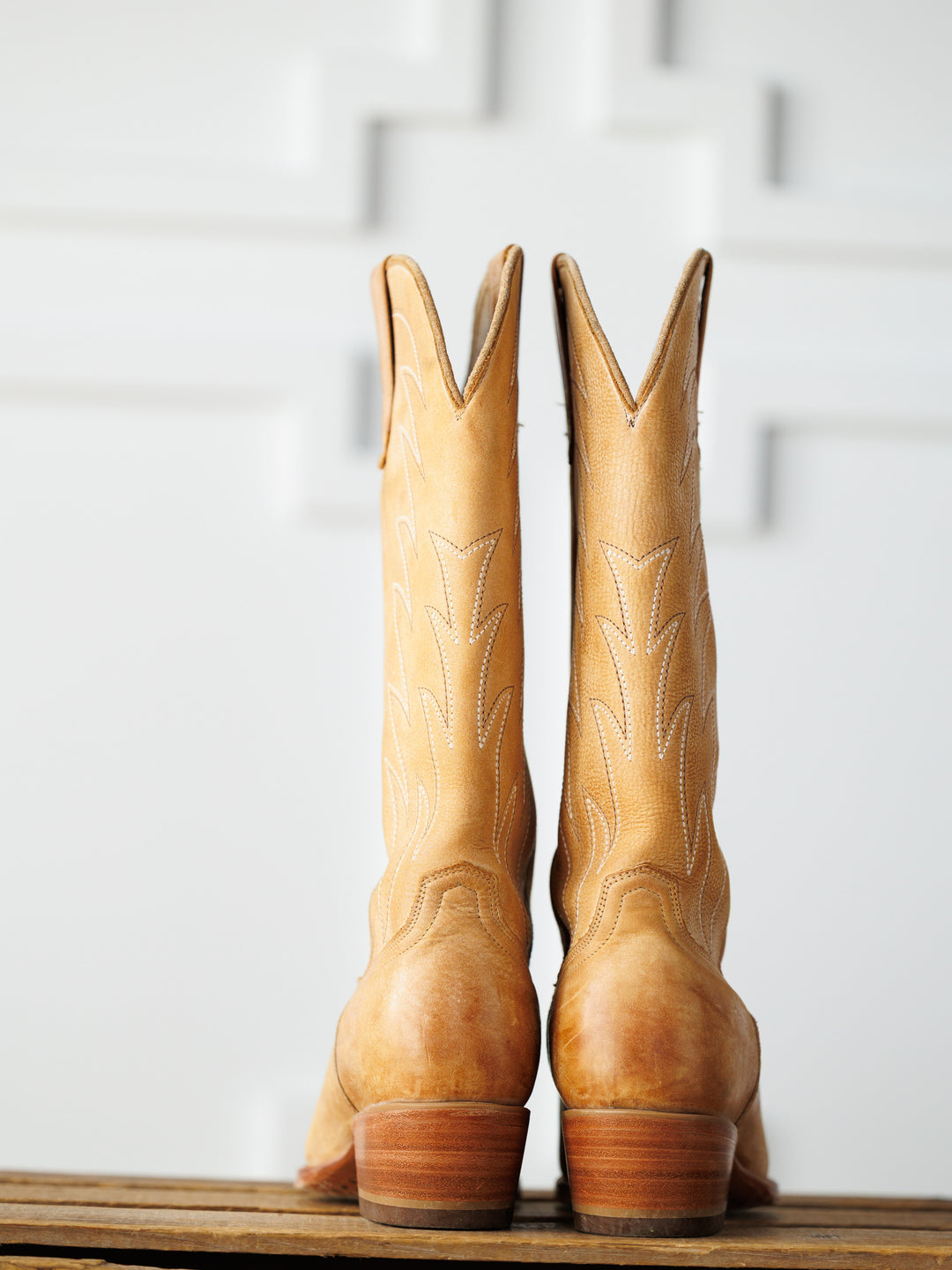The Carmencita by Savannah Sevens western life{&}style: a pair of tall, saddle tan leather western boots with decorative stitching stands side by side on a wooden surface against a white-textured background.