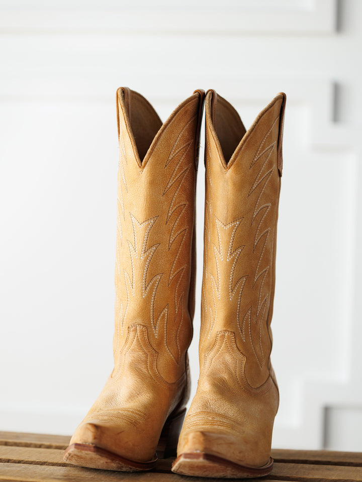 The Carmencita boots by Savannah Sevens western life{&}style, crafted in saddle tan leather with decorative stitching, stand side by side on a wooden surface against a white background. Boots made by Black Star. 