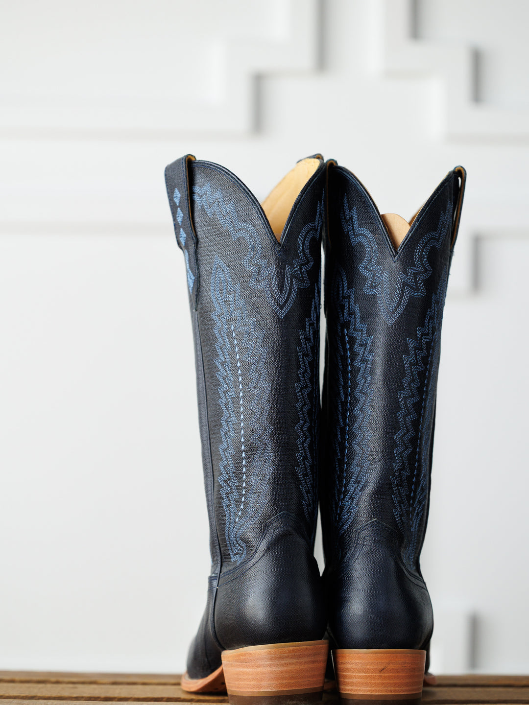 A pair of tall midnight navy leather "The Pasadena" boots by Savannah Sevens western life{&}style, featuring decorative stitching, stands side by side on a wooden surface against a white geometric-patterned background. Boots made by Blackstar.