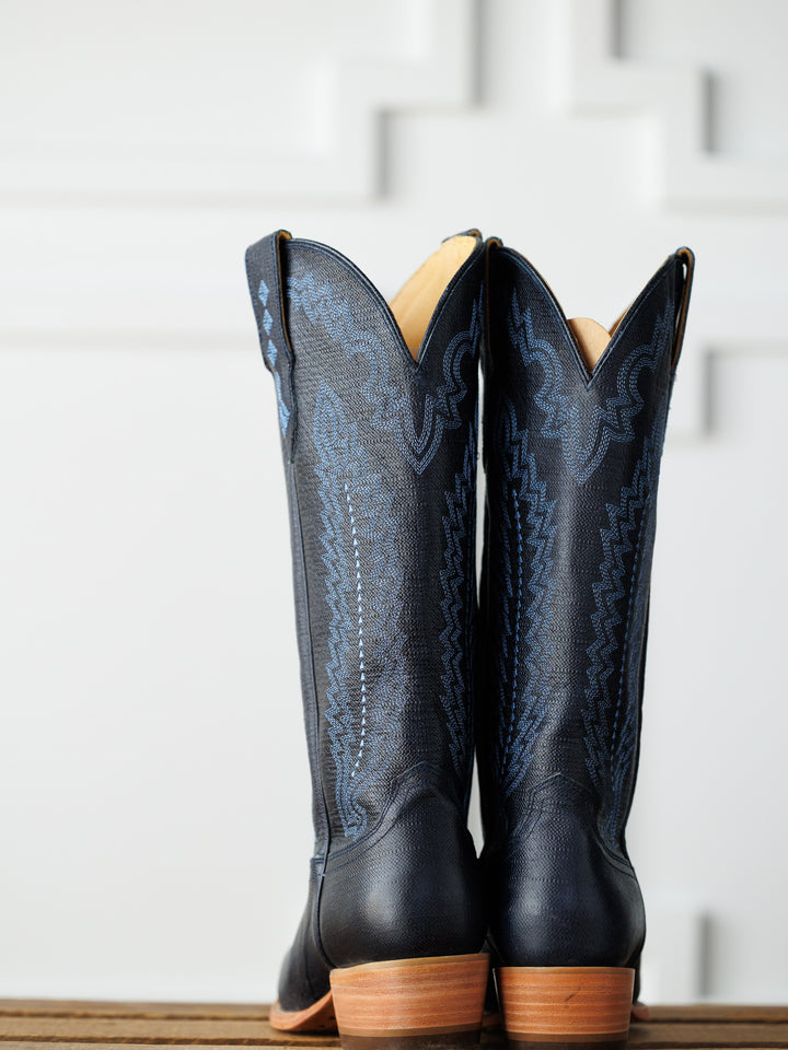 A pair of tall midnight navy leather "The Pasadena" boots by Savannah Sevens western life{&}style, featuring decorative stitching, stands side by side on a wooden surface against a white geometric-patterned background. Boots made by Blackstar.