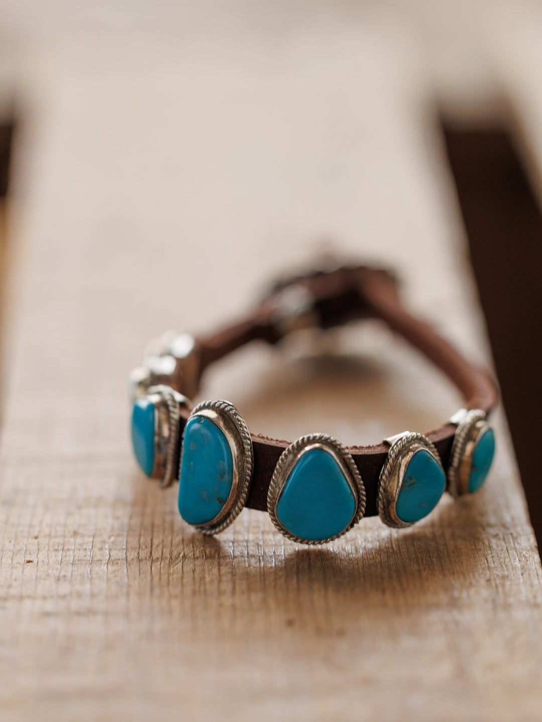 The Aguila Bracelet by Savannah Sevens western life{&}style features five genuine turquoise stones set in sterling silver on leather, displayed on a wooden surface with a blurred background.