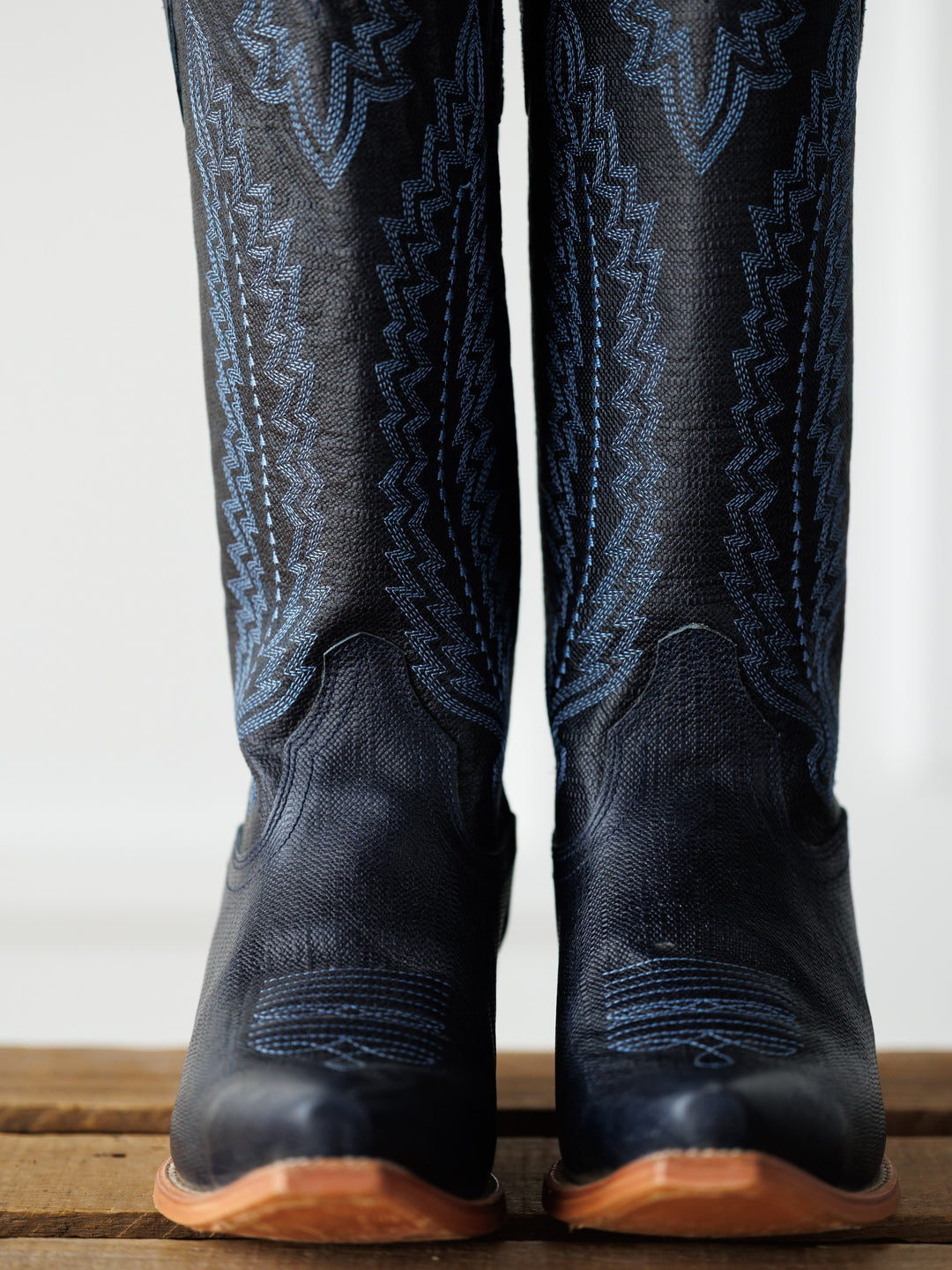 A close-up of The Pasadena boots by Savannah Sevens western life{&}style showcases black leather with blue stitching, standing upright on wood. These western snip toe boots feature sleek midnight navy accents.