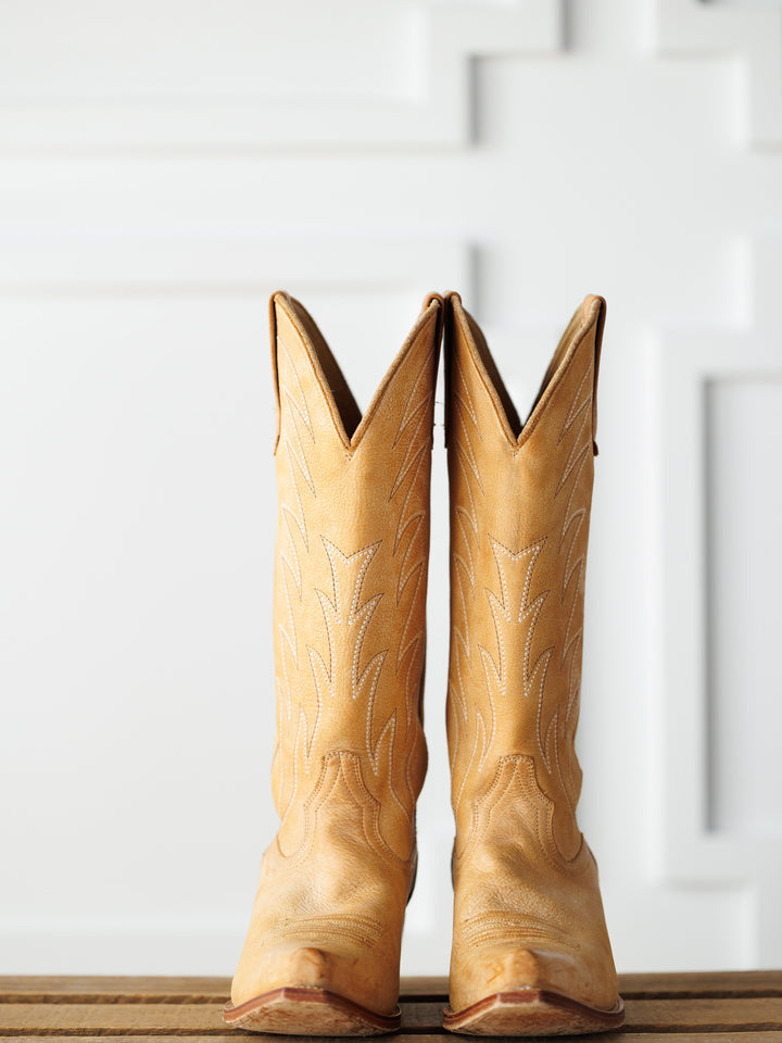The Carmencita by Savannah Sevens western life{&}style: tall saddle tan leather western boots with decorative stitching, displayed upright on a wooden surface against a white paneled backdrop. Boots made by Black Star. 