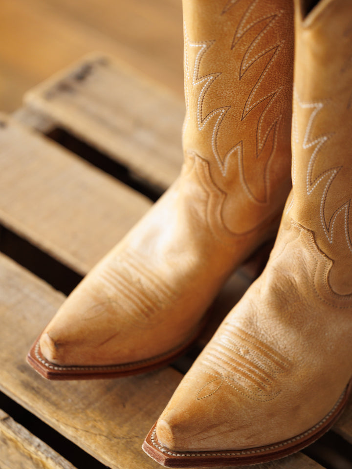 A close-up of The Carmencita saddle tan leather western boots by Savannah Sevens western life{&}style, featuring stitched designs, standing on a wooden pallet.