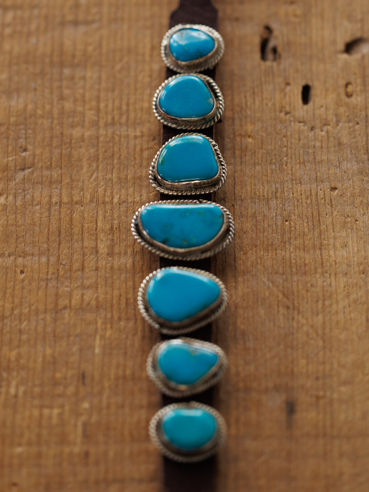 The Aguila Bracelet by Savannah Sevens western life{&}style features seven genuine turquoise stones set in sterling silver bezels on leather, shown on a wooden surface.