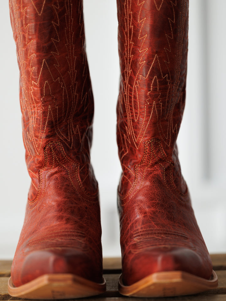 A close-up of The Pueblo snip toe boots by Savannah Sevens western life{&}style, crafted in cranberry distressed leather with intricate stitching, standing side by side on a wooden surface against a lightly blurred background. Boots made by Blackstar.