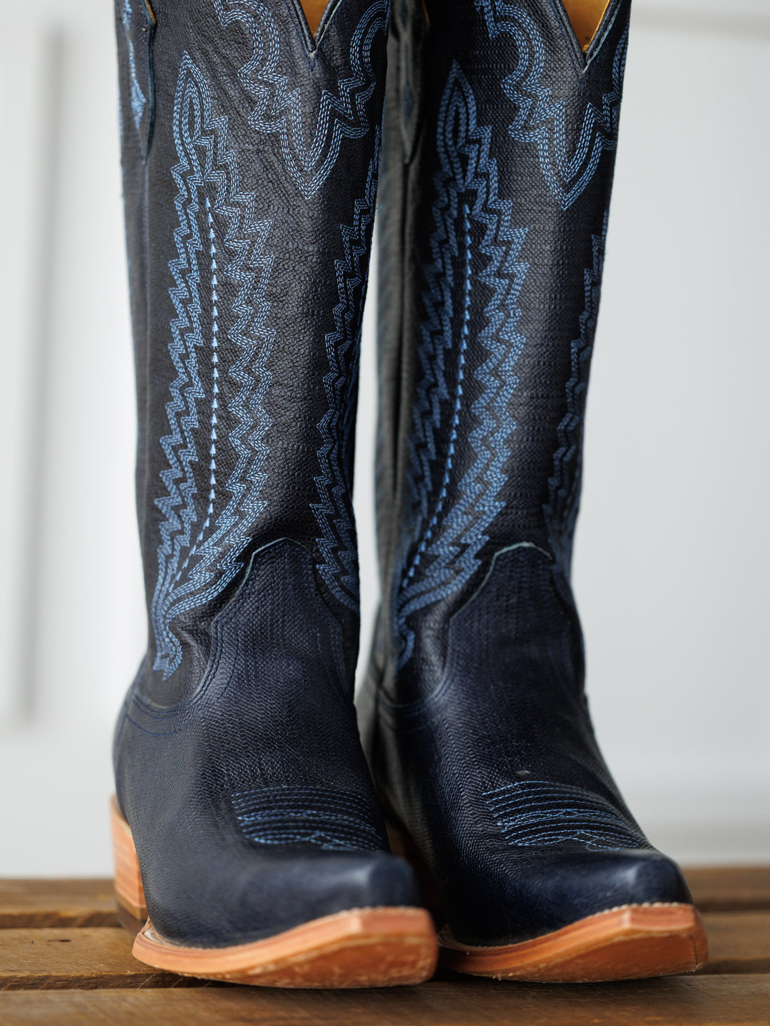 A close-up of The Pasadena boots by Savannah Sevens western life{&}style in dark blue with light blue stitching and tan leather soles, standing on a wooden surface. Boots made by Blackstar.