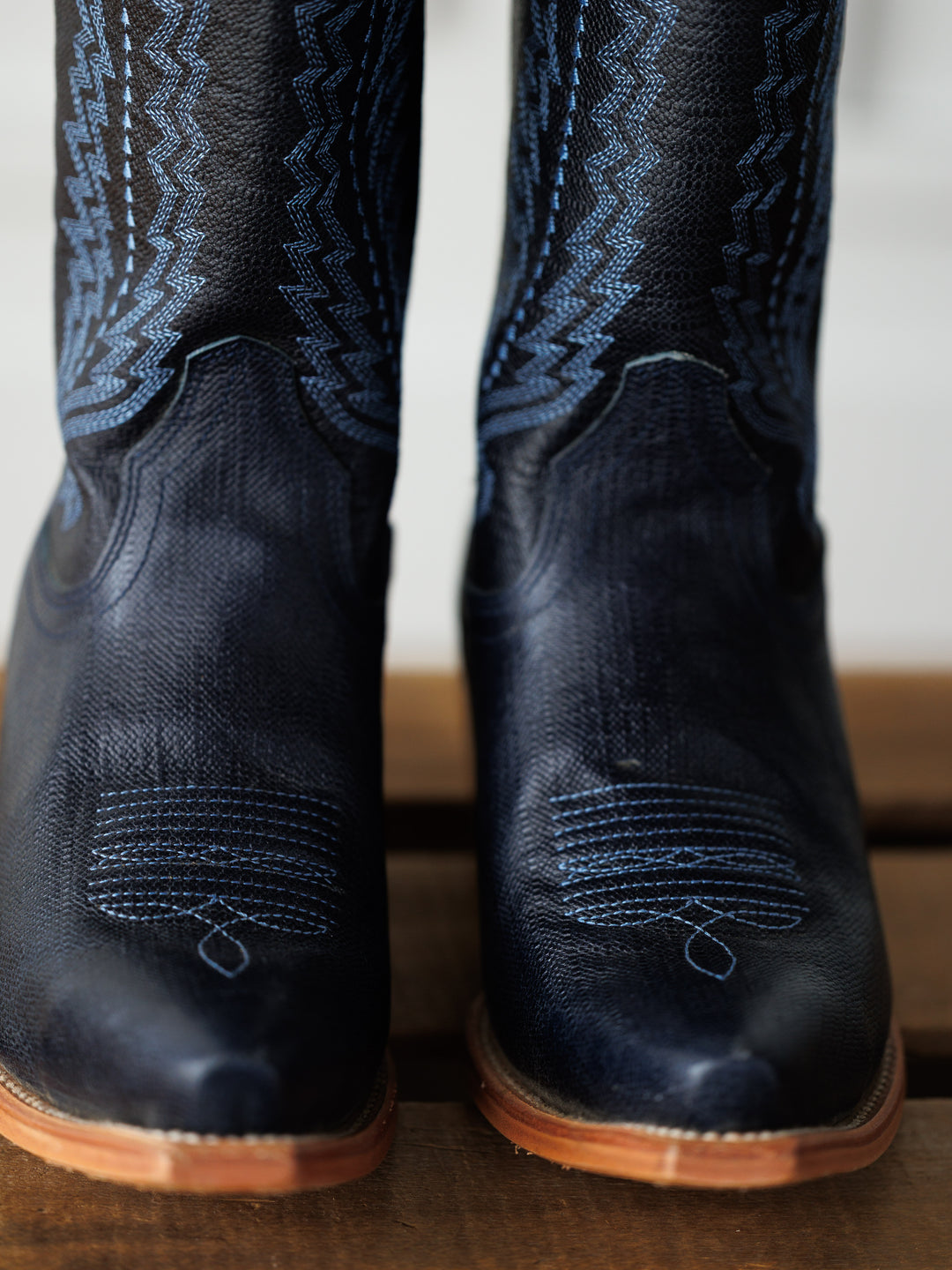 Close-up of The Pasadena boots by Savannah Sevens western life{&}style in dark blue with decorative blue stitching and light brown soles, standing on a wooden surface. Boots made by Black Star. 