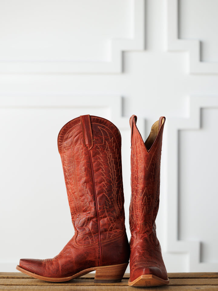 The Pueblo by Savannah Sevens western life{&}style, tall cranberry leather snip toe western boots with decorative stitching, stands on a wooden surface against a white geometric-patterned wall. Boots made by Black Star. 