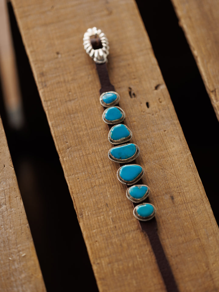 The Aguila Bracelet by Savannah Sevens western life{&}style features six genuine turquoise stones set in sterling silver on leather, arranged in a row and displayed on a wooden surface.