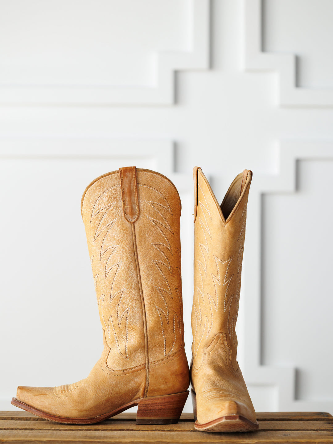 The Carmencita western boots by Savannah Sevens western life{&}style, in saddle tan leather with decorative stitching—one upright, one on its side—are displayed on a wooden surface against a white patterned background.