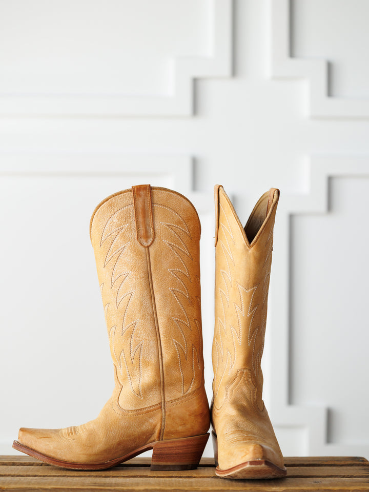The Carmencita western boots by Savannah Sevens western life{&}style, in saddle tan leather with decorative stitching—one upright, one on its side—are displayed on a wooden surface against a white patterned background.