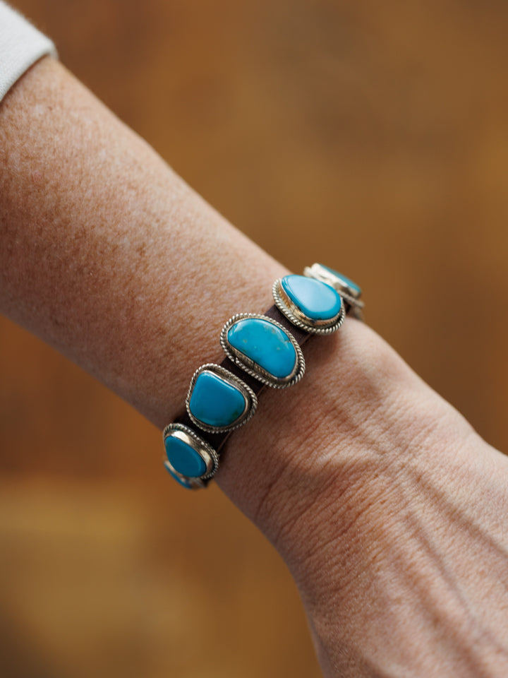 A close-up shows a wrist wearing the Aguila Bracelet by Savannah Sevens western life{&}style, a sterling silver piece set with five irregular genuine turquoise stones, against a soft brown background.