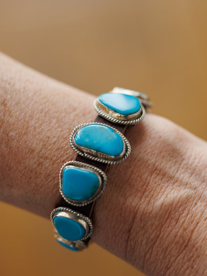 A close-up of a wrist wearing the Aguila Bracelet by Savannah Sevens western life{&}style, crafted in sterling silver with five large, irregular genuine turquoise stones set in decorative bezels against a softly blurred background.