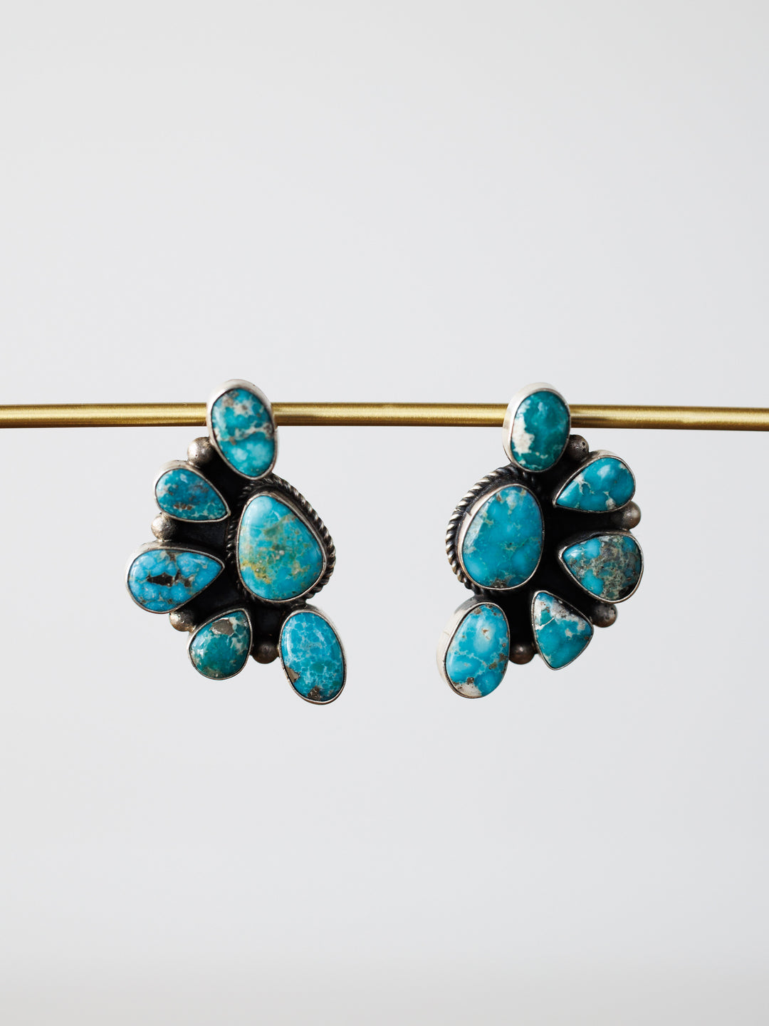 The Watseka Earrings by Savannah Sevens western life{&}style are Native American handcrafted sterling silver earrings featuring turquoise stones in a fan shape, displayed hanging from a gold rod on a plain white background.
