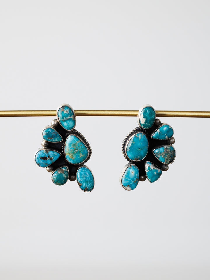 The Watseka Earrings by Savannah Sevens western life{&}style are Native American handcrafted sterling silver earrings featuring turquoise stones in a fan shape, displayed hanging from a gold rod on a plain white background.