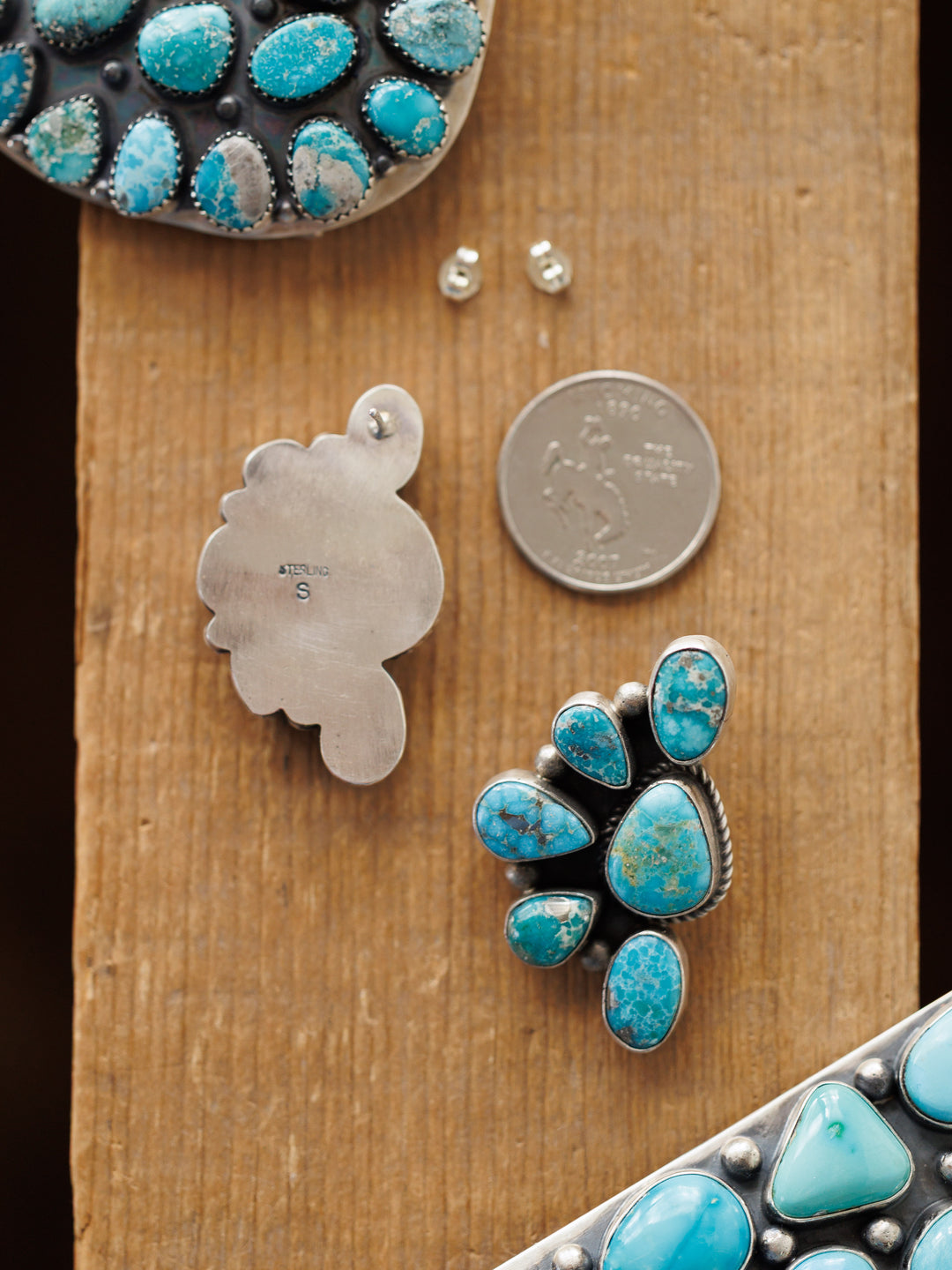 The Watseka Earrings by Savannah Sevens western life{&}style, along with a silver pendant, earring backs, and a U.S. quarter, rest on a wooden surface. The Native American handcrafted turquoise pieces feature unique cluster designs set in silver.