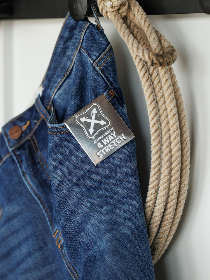 A pair of The Rhett High Rise Relaxed Straight Leg Jeans by Savannah Sevens western life{&}style hangs next to a coiled rope, featuring a waistband tag that reads 4 WAY STRETCH.