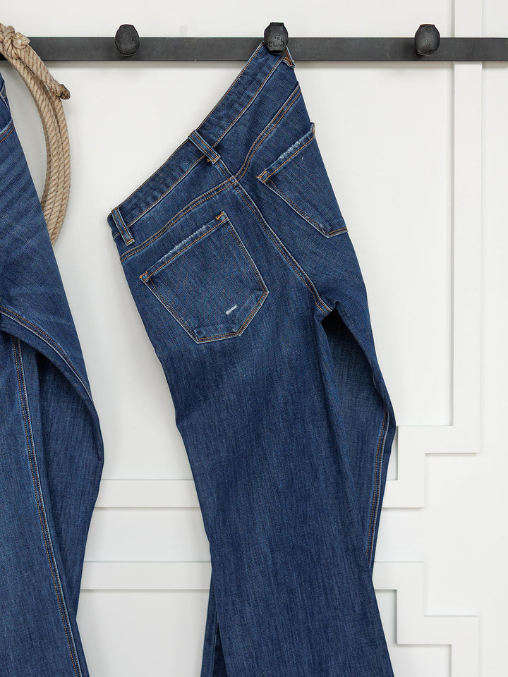 The Rhett High Rise Relaxed Straight Leg Jean by Savannah Sevens western life{&}style hangs by the waistband on a black metal hook against a white paneled wall, with another relaxed straight leg jean partially visible to the left.