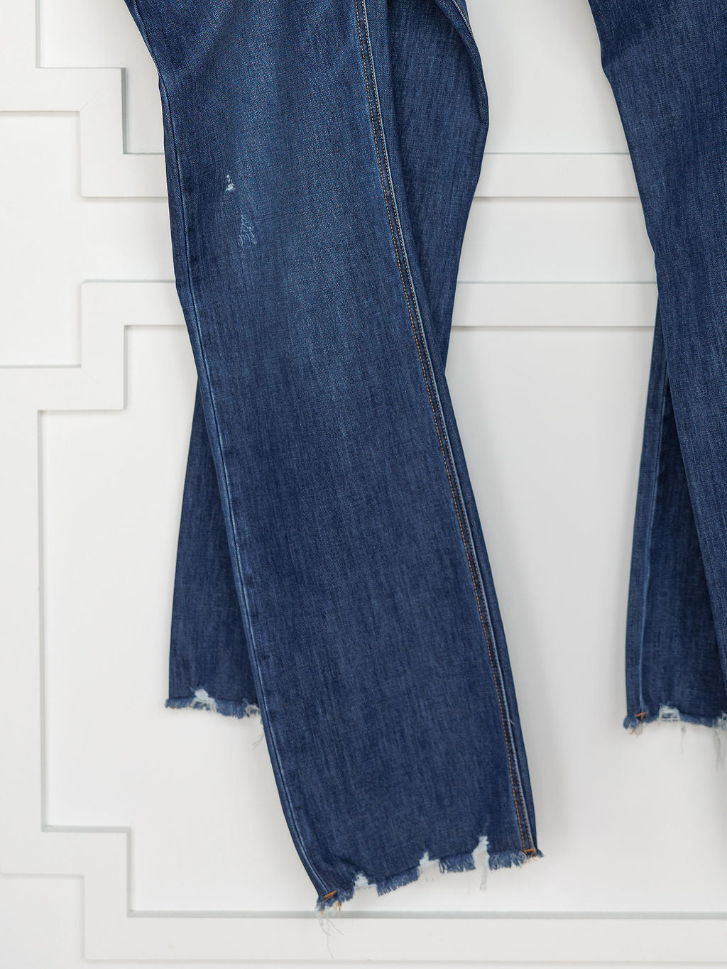 A close-up of The Rhett High Rise Relaxed Straight Leg Jean by Savannah Sevens western life{&}style, featuring blue denim with raw frayed hems and light distressing at the knees, displayed against a white textured wall.