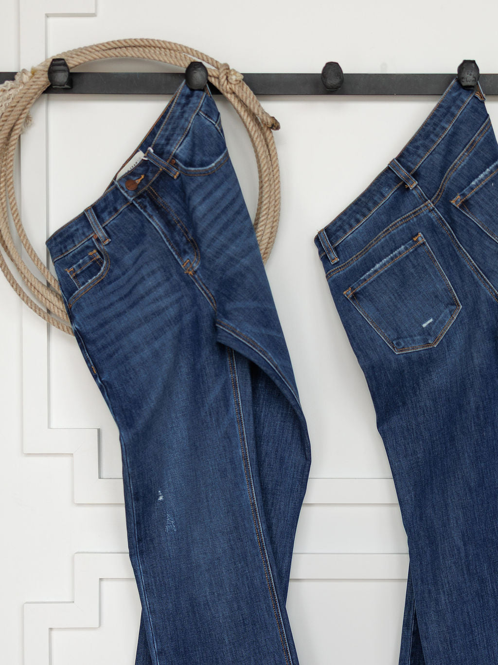 Two pairs of The Rhett High Rise Relaxed Straight Leg Jeans by Savannah Sevens western life{&}style hang from black hooks on a white wall, with a coiled rope beside them for a rustic, western accent in the minimalist space.
