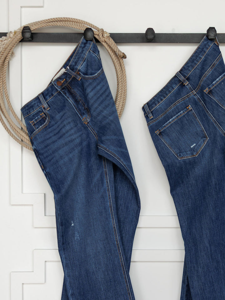Two pairs of The Rhett High Rise Relaxed Straight Leg Jeans by Savannah Sevens western life{&}style hang from black hooks on a white wall, with a coiled rope beside them for a rustic, western accent in the minimalist space.