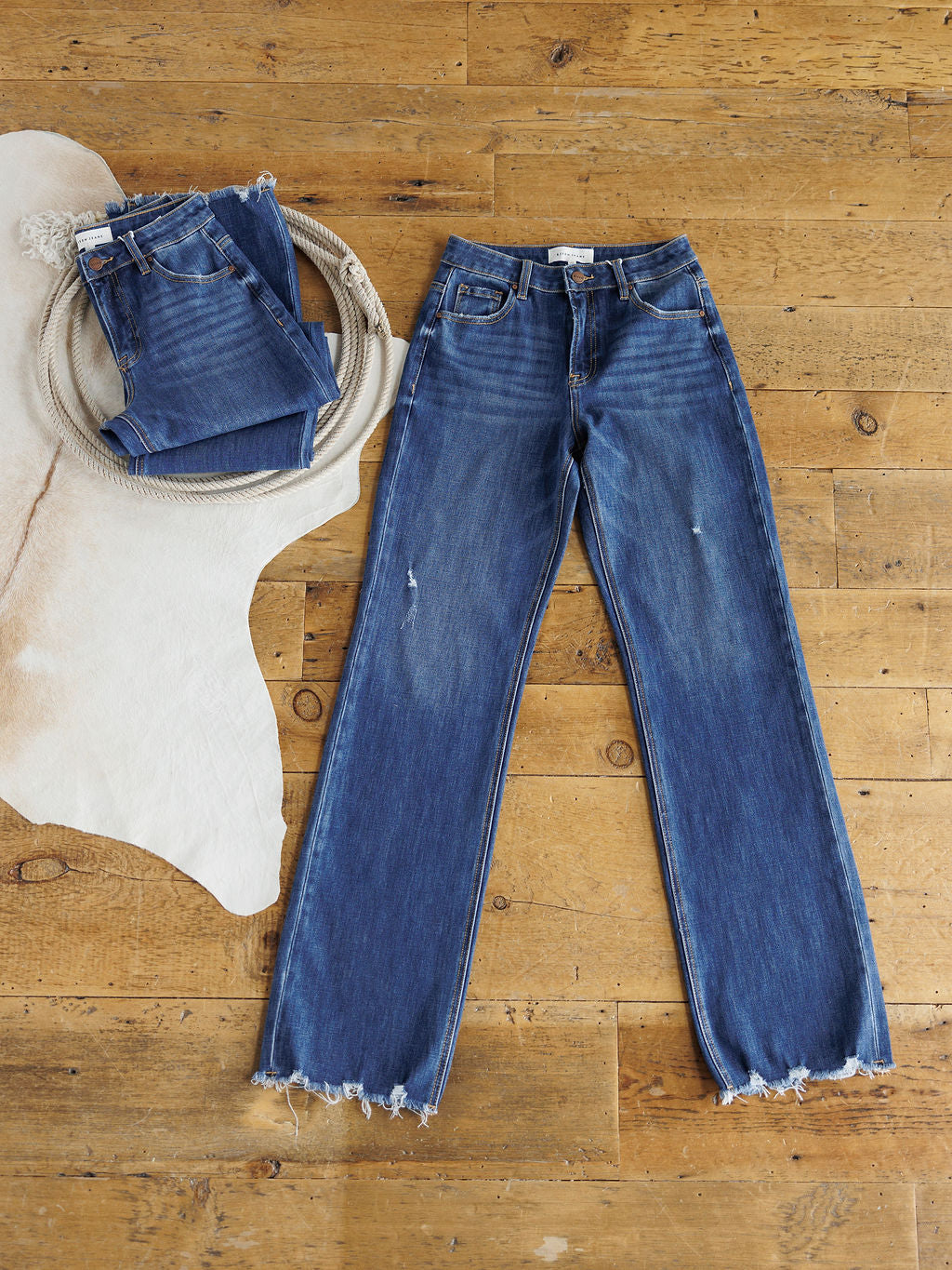 A pair of blue jeans with raw, frayed hems lies flat on a wooden floor near a cowhide rug and coiled rope, with another pair—the Rhett High Rise Relaxed Straight Leg Jean by Savannah Sevens western life{&}style—folded neatly on top.