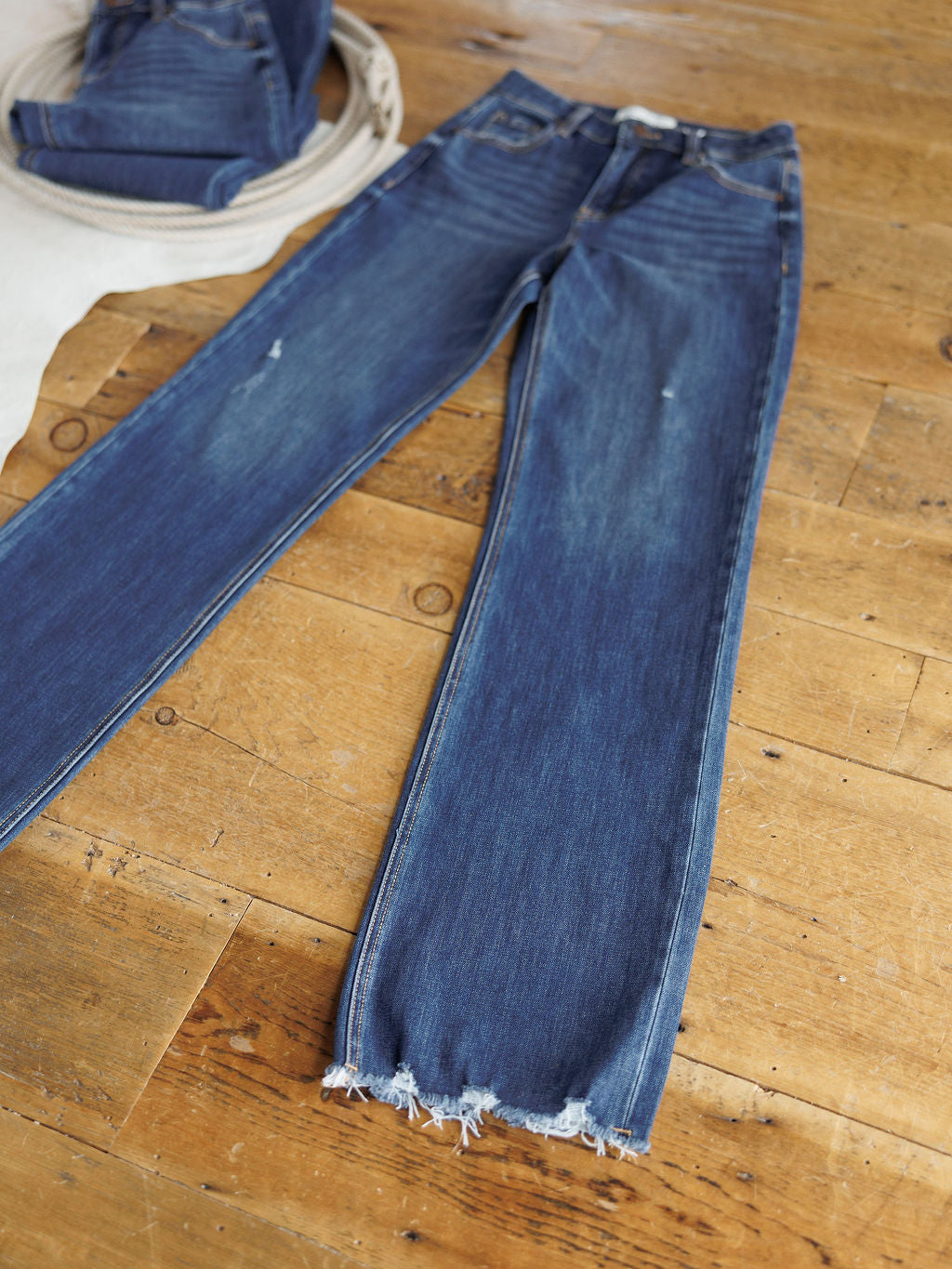 The Rhett High Rise Relaxed Straight Leg Jean from Savannah Sevens western life{&}style in blue denim with slight distressing and frayed hems is laid flat on a wooden floor, with another folded pair partially visible in the background.