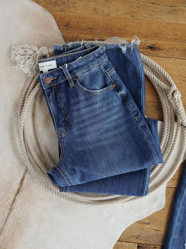 The Rhett High Rise Relaxed Straight Leg Jean by Savannah Sevens western life{&}style, folded with a frayed hem, rests atop a coiled rope and light animal hide on a wooden floor.