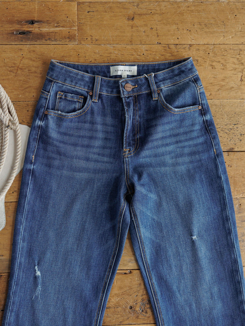 The Rhett High Rise Relaxed Straight Leg Jean by Savannah Sevens western life{&}style, in blue with subtle fading and slight distressing, lays flat on a wooden floor with a coiled rope and white fabric at the edge of the image.
