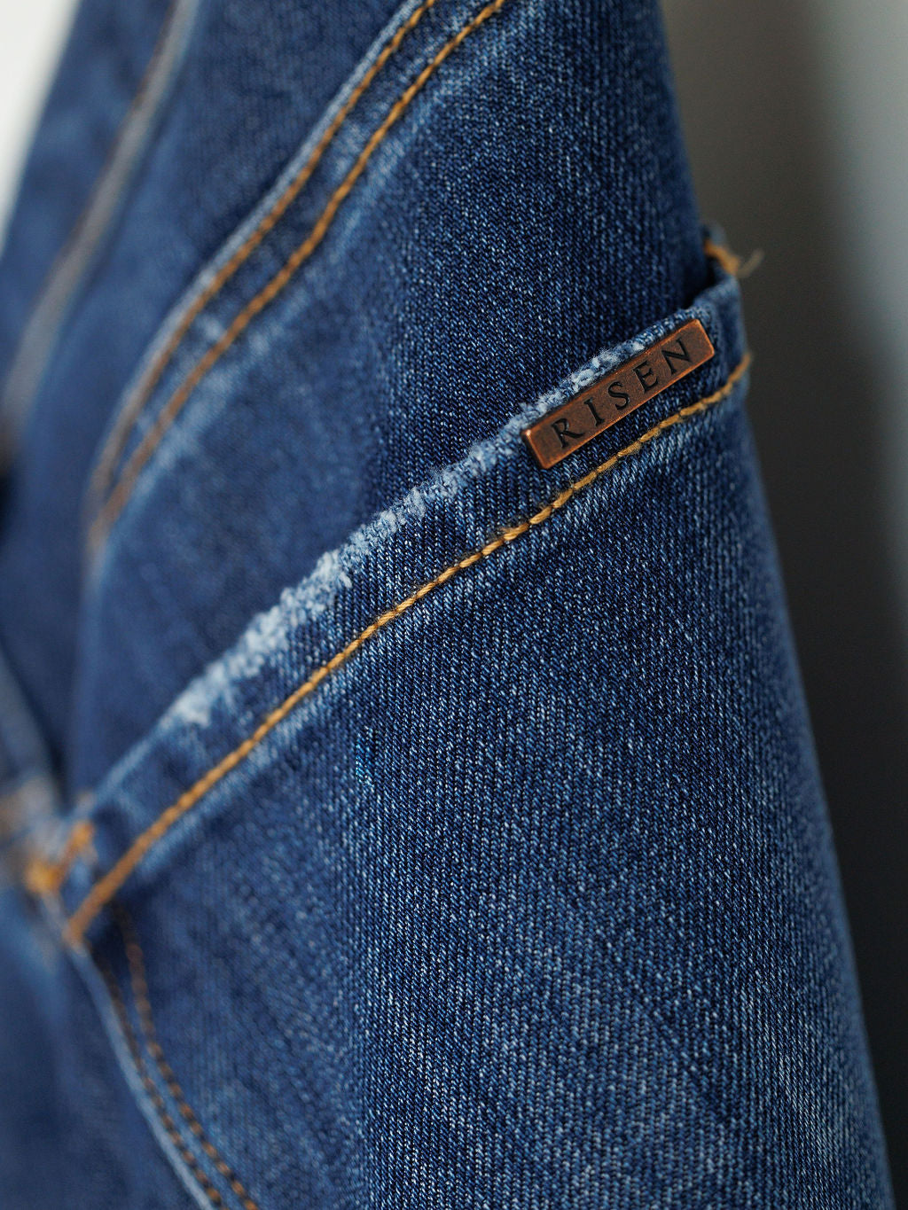 Close-up of blue denim The Rhett High Rise Relaxed Straight Leg Jean by Savannah Sevens western life{&}style, featuring orange stitching and a small metallic tag near the seam. These jeans highlight quality craftsmanship and classic style.