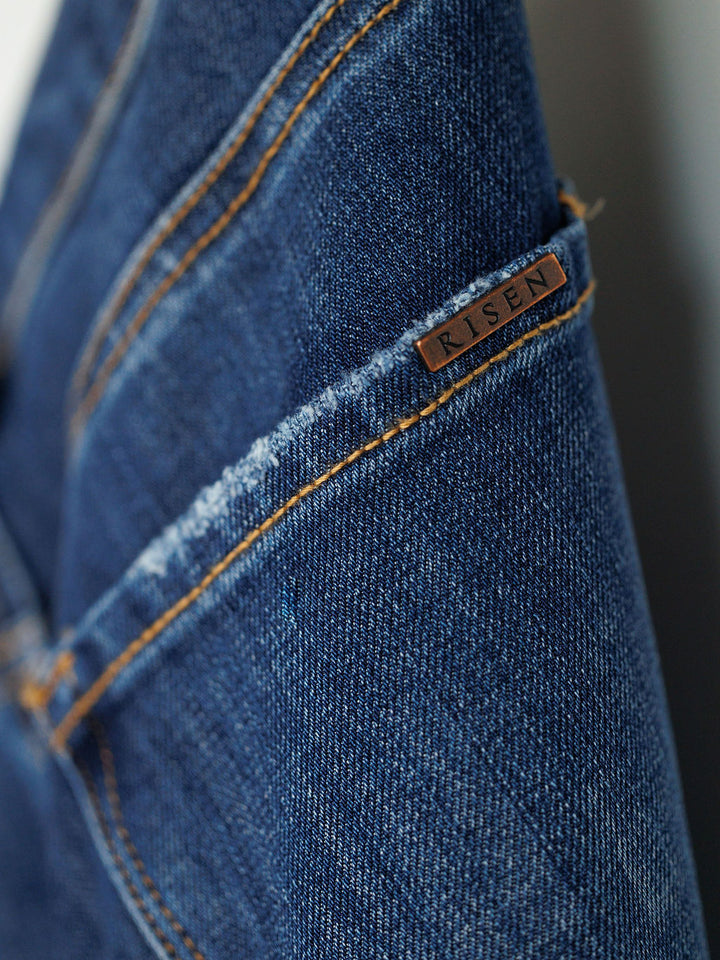 Close-up of blue denim The Rhett High Rise Relaxed Straight Leg Jean by Savannah Sevens western life{&}style, featuring orange stitching and a small metallic tag near the seam. These jeans highlight quality craftsmanship and classic style.