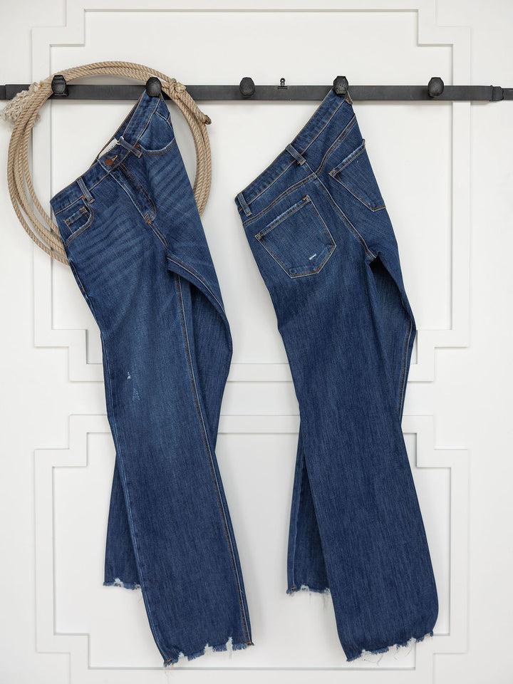 Two pairs of The Rhett High Rise Relaxed Straight Leg Jeans by Savannah Sevens western life{&}style hang on a black rod against a white wall—one suspended by a rope, the other hanging directly on the rod, both featuring frayed hems.