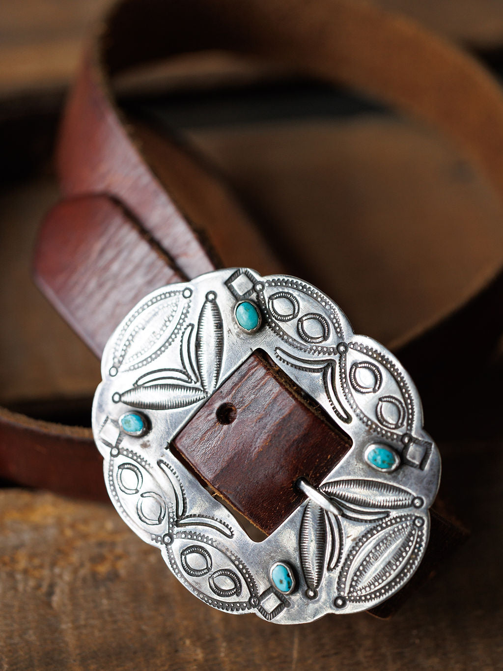 A close-up of the Ruidoso Belt by Savannah Sevens western life{&}style, showcasing its brown leather, engraved sterling silver Navajo buckle, and turquoise stones, displayed on a wooden surface.
