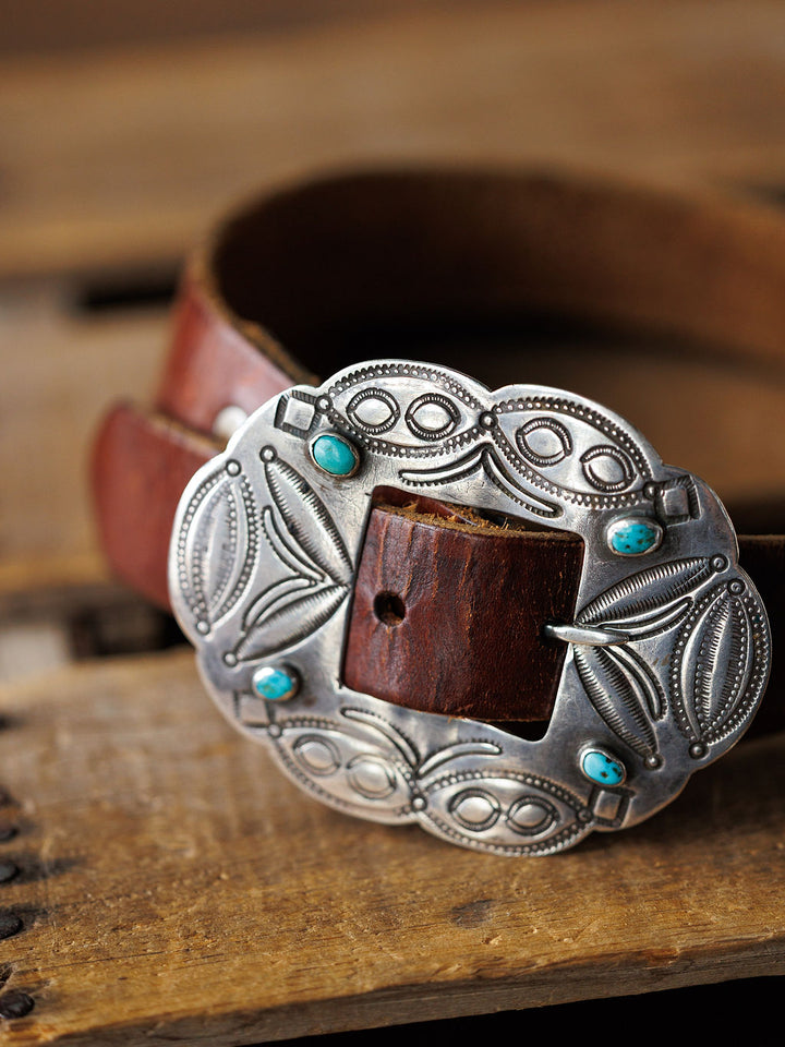 A close-up of the Ruidoso Belt by Savannah Sevens western life{&}style, featuring a brown leather strap and an ornate sterling silver Navajo buckle set with four turquoise stones, displayed on a wooden surface.