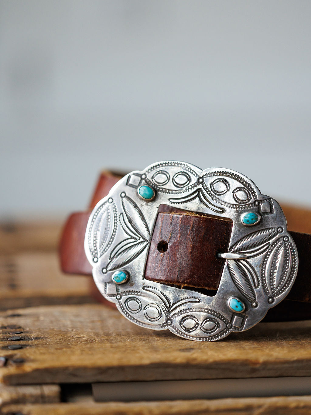 The Ruidoso Belt by Savannah Sevens western life{&}style is a brown leather belt with a Navajo-style engraved sterling silver buckle and four turquoise stones, displayed on a wooden surface.