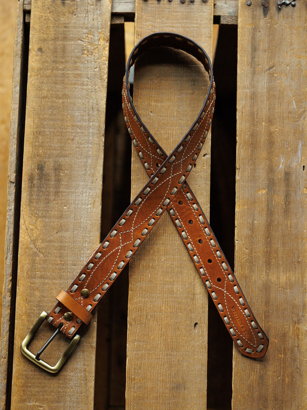 The Barlow Belt by Savannah Sevens western life{&}style features buck stitch detail and metal eyelets, coiled on a wooden pallet with its brass buckle visible in the lower left corner.