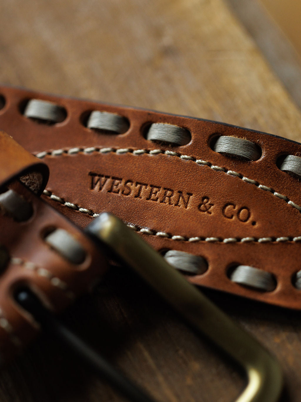 Close-up of the Barlow Belt by Savannah Sevens western life{&}style, featuring decorative stitching and buck stitch on brown leather with an interchangeable metal buckle, shown resting on a wooden surface.