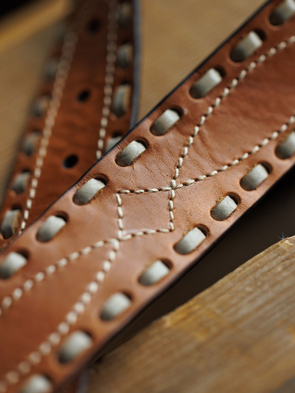 Close-up of the Barlow Belt by Savannah Sevens western life{&}style, featuring decorative white stitching and gray lacing through large holes with buck stitch detail, displayed on a wooden surface.