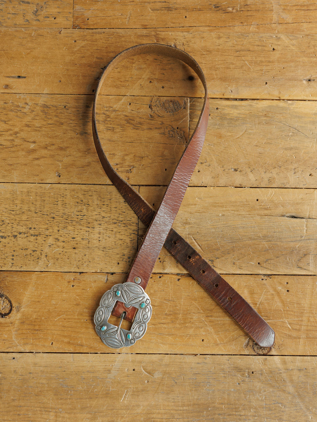 The Ruidoso Belt by Savannah Sevens western life{&}style, featuring a large engraved sterling silver buckle and brown leather, is displayed in a loose loop on a wooden surface.